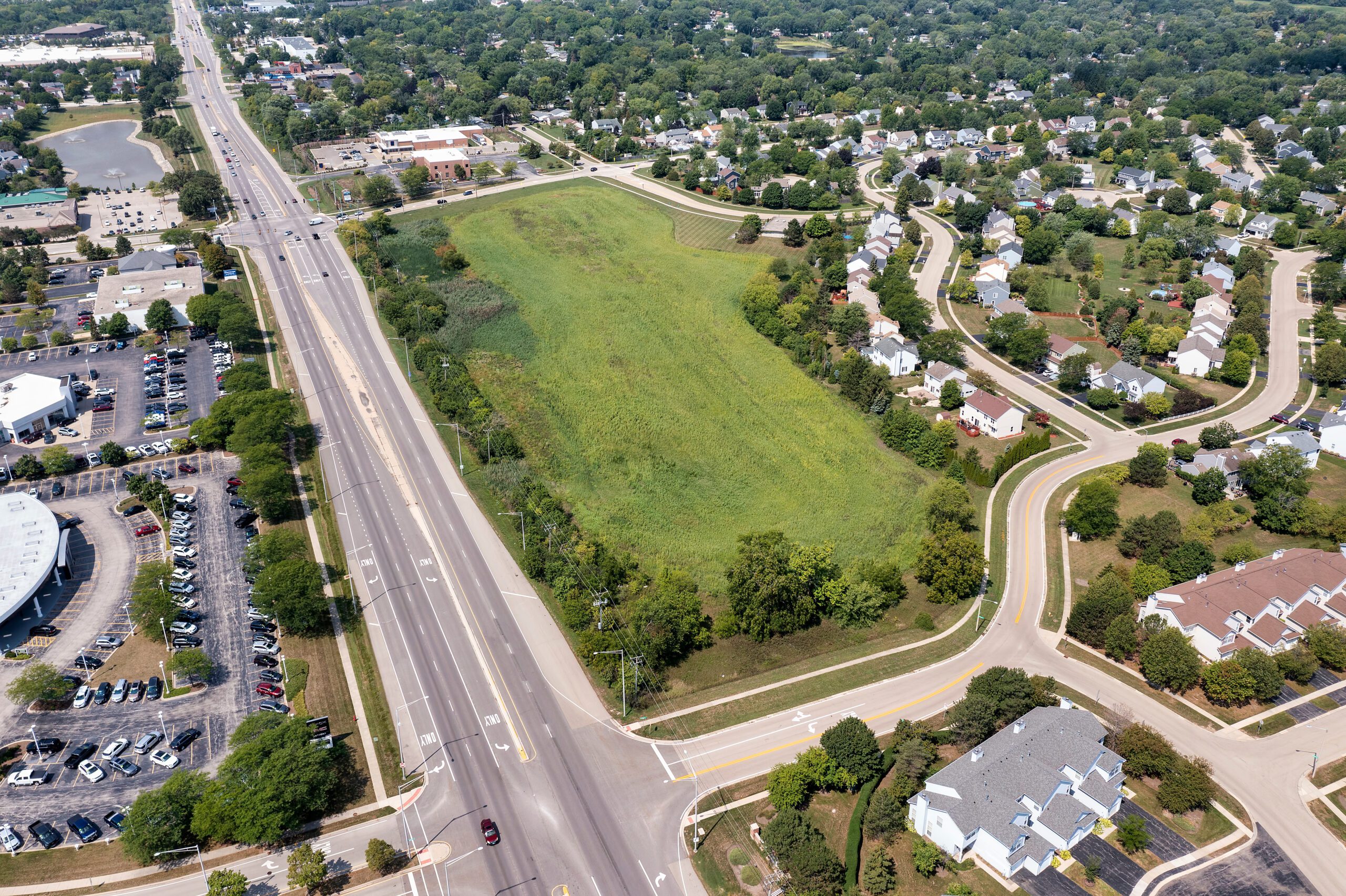 Juno all in one Services - Aerial view of a green open field in Texas, surrounded by residential houses, roads, and businesses. Cars drive on a main road and parking lots visible to the left. Trees and sidewalks line the well-maintained neighborhood streets.